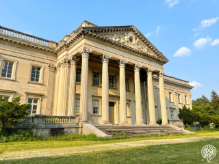 View of Lynnewood Hall's front portico and columns. The detailed pediment with multiple human figures is atop tall Corinthian columns. The foreground is bright green grass and the sky is soft blue with a few clouds.