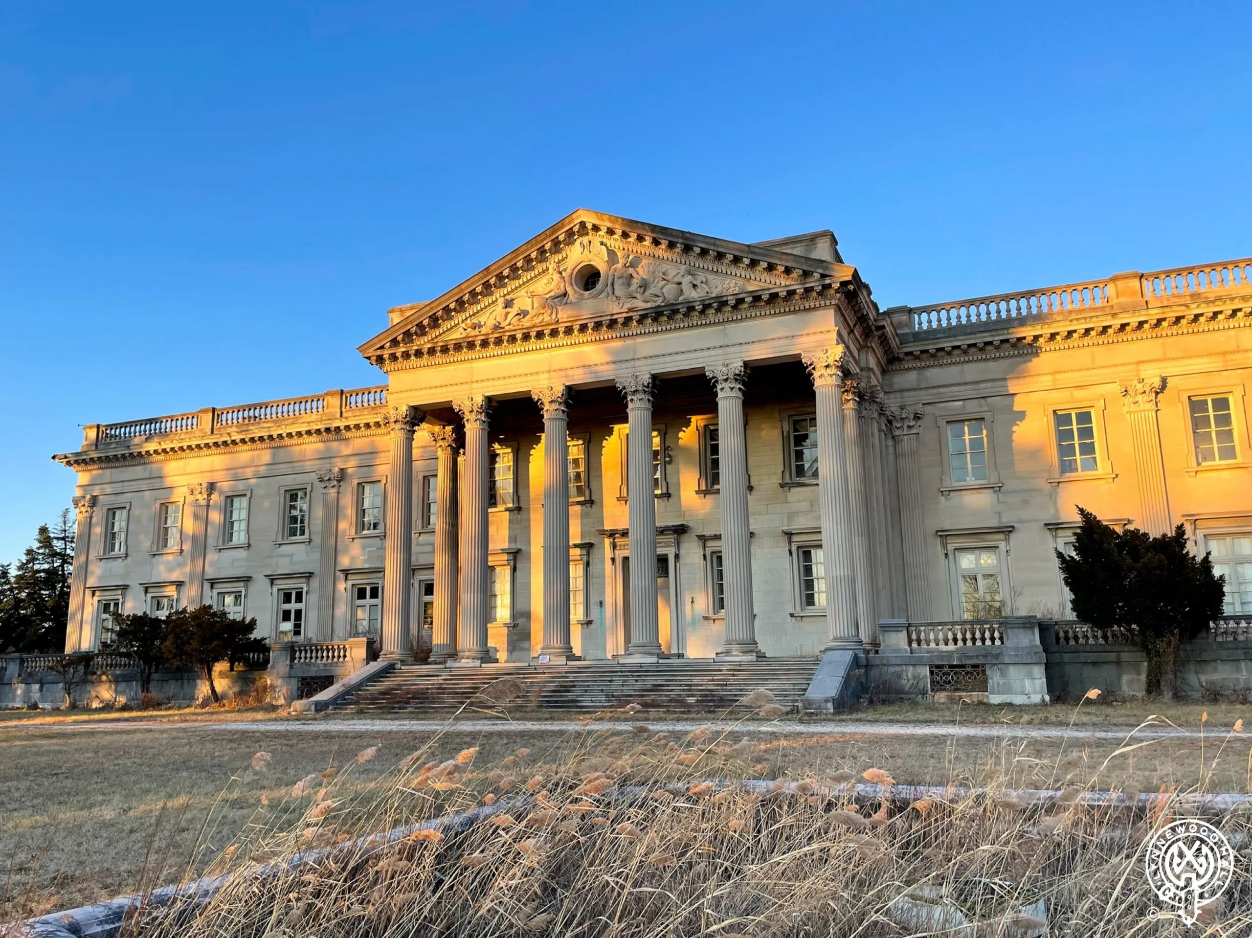 View of front elevation of Lynnewood Hall at early sunset. The stone facade is reflecting the golden sun. In the foreground is tall brown grass and the sky is bright blue.